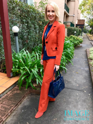 Woman wearing an orange suit and blue top stands near a garden.