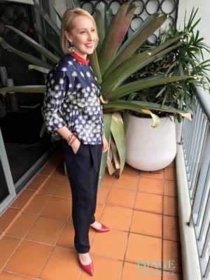 Woman stands on a balcony wearing a red blouse and shoes, and navy pants and jacket.