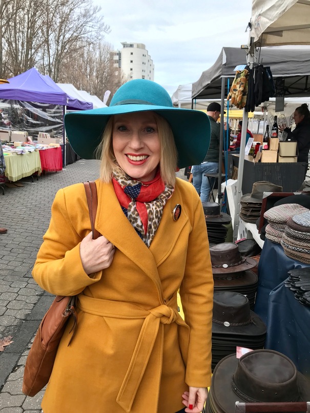 Woman at an open air market wearing golden yellow coloured coat, teal felt hat and animal print scarf.