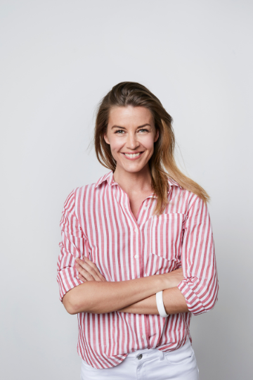 Smiling woman wears a red and white stripe blouse with white jeans. She has her arms crossed.