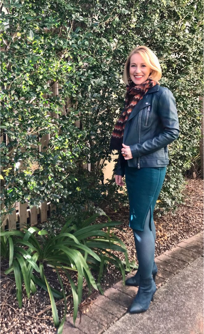 A women is standing in a garden. She is wearing various layering clothes for the cool autumn day. These items include a leather jacket, teal coloured hosiery and a woollen scarf.