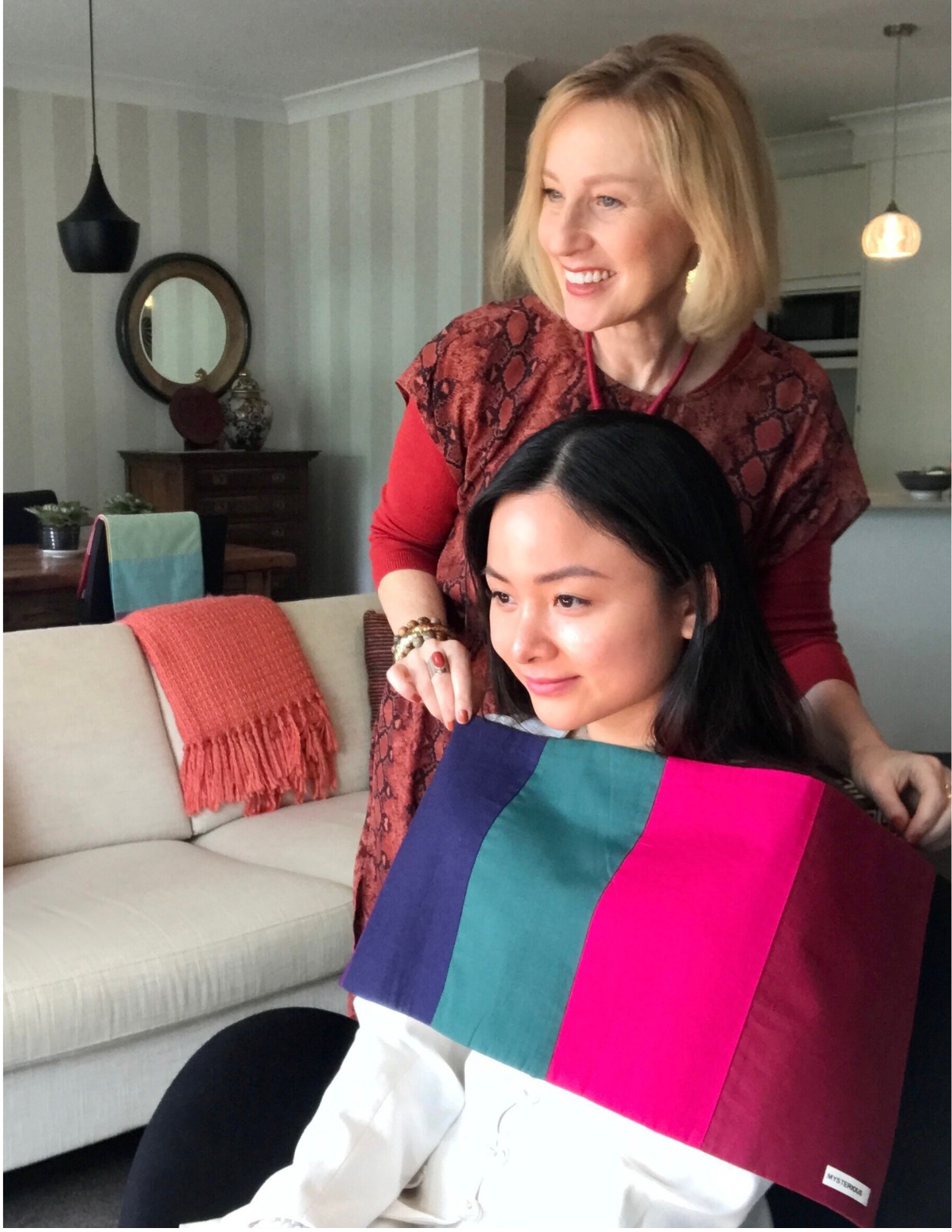 A woman sits in a chair. There is a lady standing behind her draping a range of coloured fabrics under her chin. Both women are smiling because the colours look beautiful on her. The woman in the chair is having a colour analysis.