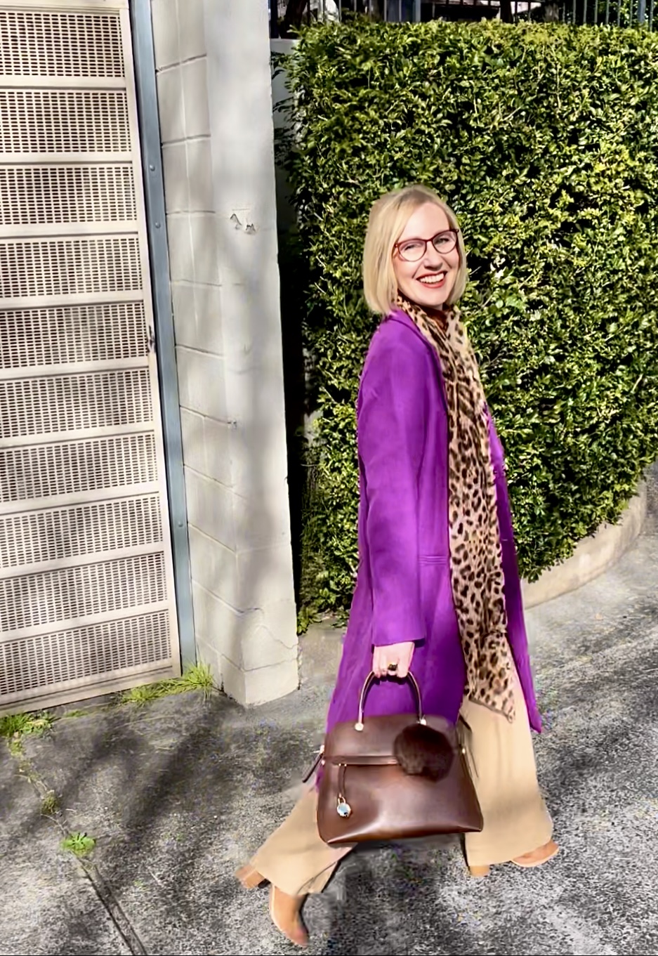 A woman is walking. She is wearing a long purple coat and a leopard print scarf. Her handbag is made from brown leather. 