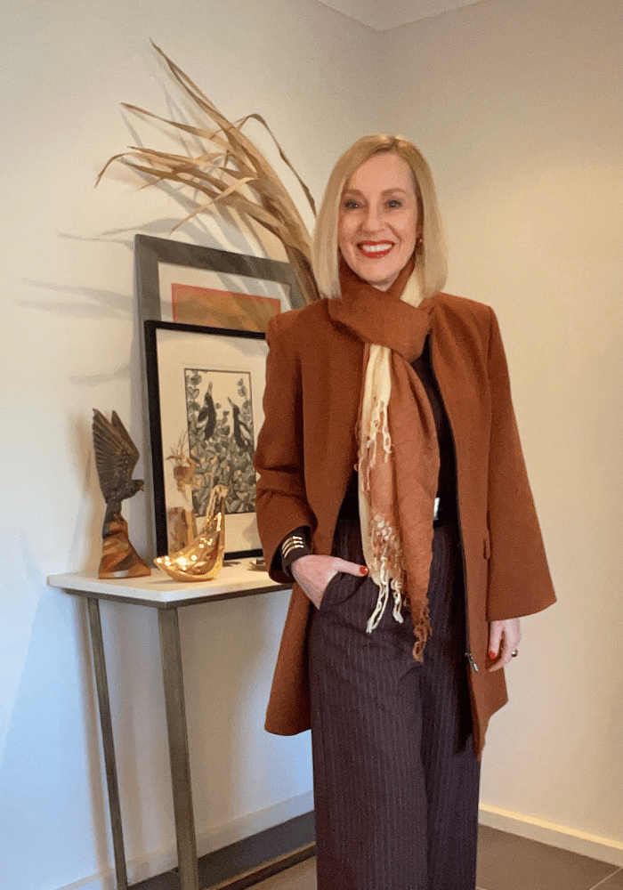 A woman is smiling. Her right hand is in the pocket of her dark brown pants. Her coat and scarf are a camel colour.