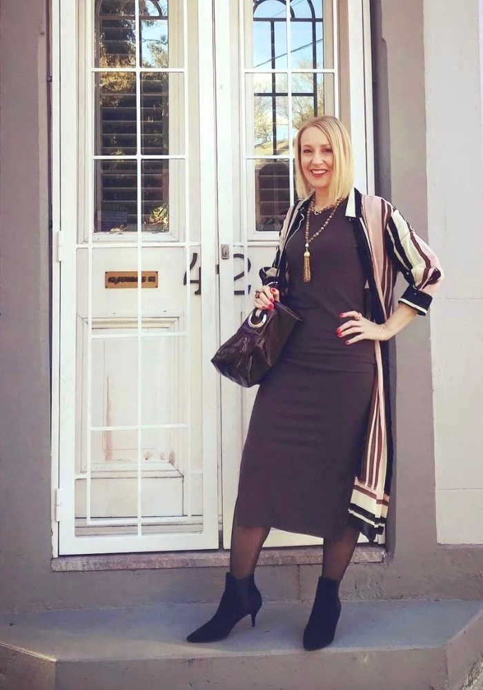 A woman is standing on a step. She is wearing a body hugging brown dress and a duster coat that features light pink stripes.