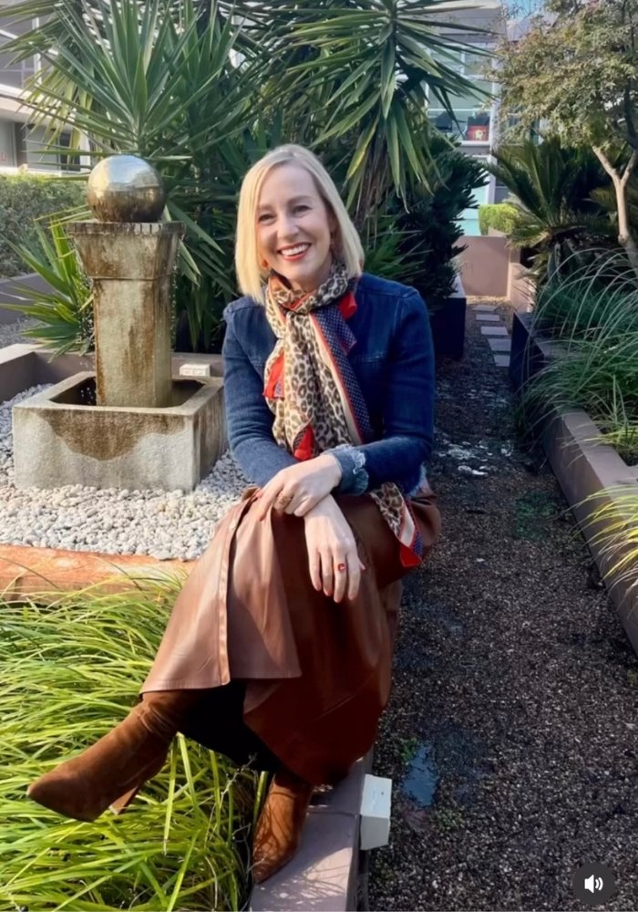 A woman is sitting on a fence. She is wearing a brown leather tiered skirt and boots. She is also wearing a navy coloured denim jacket.