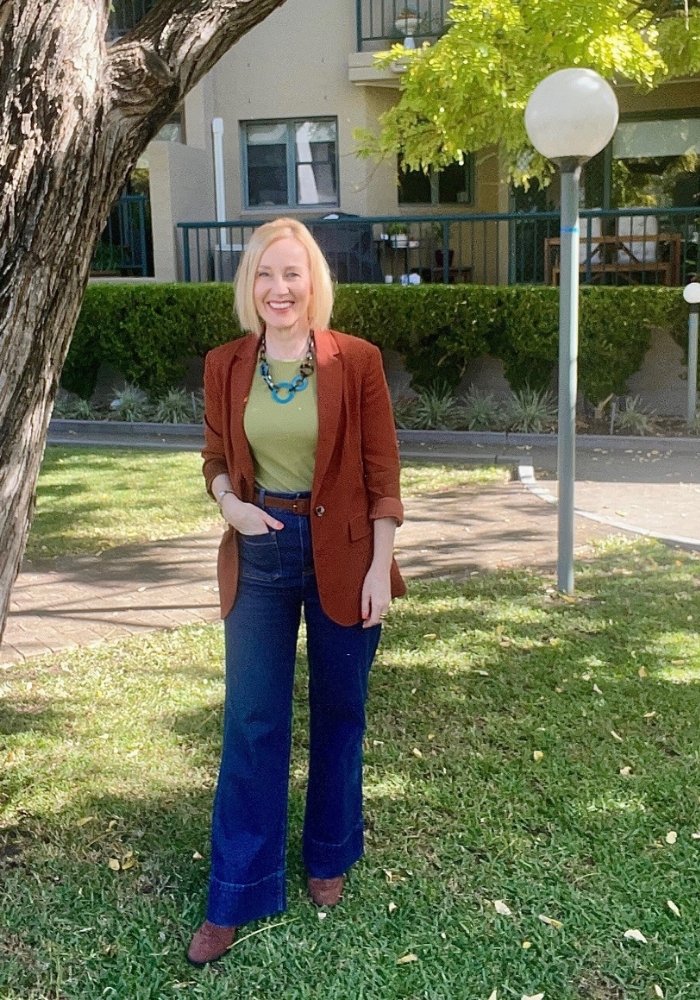 A woman stands by a tree. She is wearing a brown blazer, jeans, and light olive coloured t-shirt.