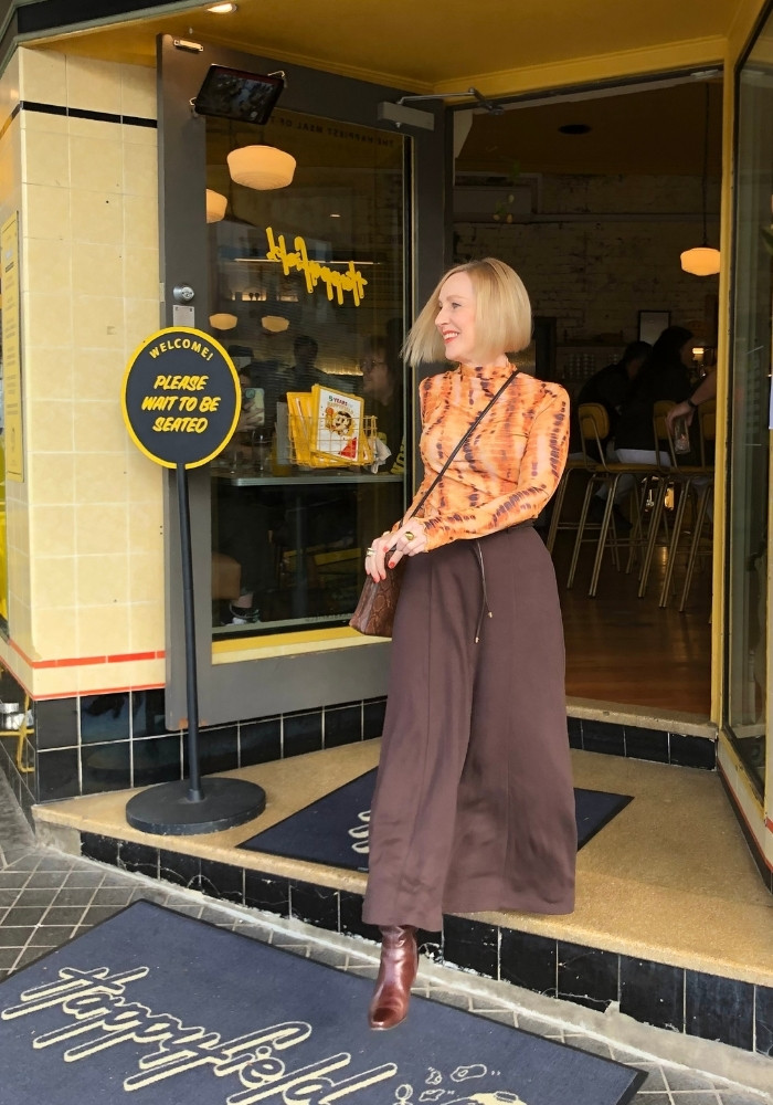 A woman is walking out of a cafe. She is wearing a brown skirt and a yellow top.