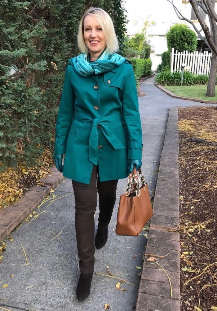 A woman is walking down a path. She is wearing dark brown jeans and boots, and a teal coloured trench coat.